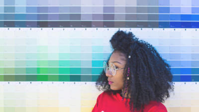A woman with large glasses and bright lipstick standing in front of a wall of color swatches.